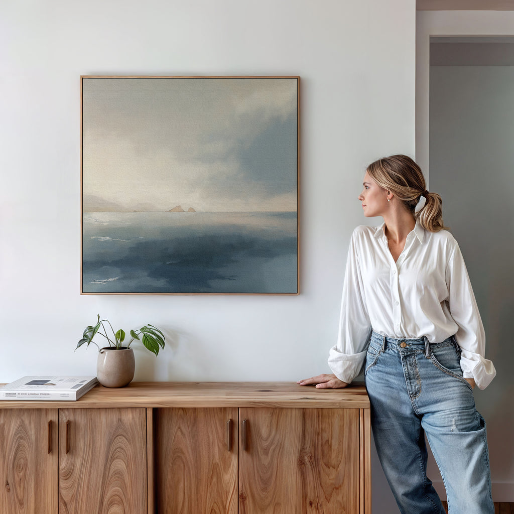 A woman in a white blouse and jeans stands by a wooden cabinet, admiring the Distant Isles Canvas Art - Square on the wall. A small plant and books sit on the cabinet, completing the serene, Nordic-inspired setting.