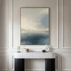 A minimalist console table with black legs and a white marble top stands against a paneled wall, decorated with modern accents and a large framed Distant Isles Canvas Art - Vertical, showcasing soft blue and beige tones of a tranquil seascape.