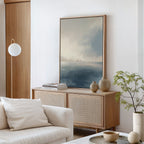 A modern living room with neutral tones showcases a wooden sideboard, ceramic vases, books, a green plant, a round pendant lamp, cushions on a white sofa, and the Distant Isles Canvas Art - Vertical as serene wall décor.