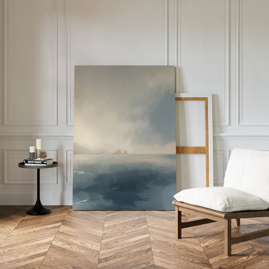 A minimalist room with wood herringbone floors, white paneled walls, a cushioned wooden chair, and a round side table set with candles and books. The Distant Isles Canvas Art - Vertical leans against the wall, bringing serene coastal calm to the space.