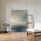 A minimalist room with wood herringbone floors, white paneled walls, a cushioned wooden chair, and a round side table set with candles and books. The Distant Isles Canvas Art - Vertical leans against the wall, bringing serene coastal calm to the space.