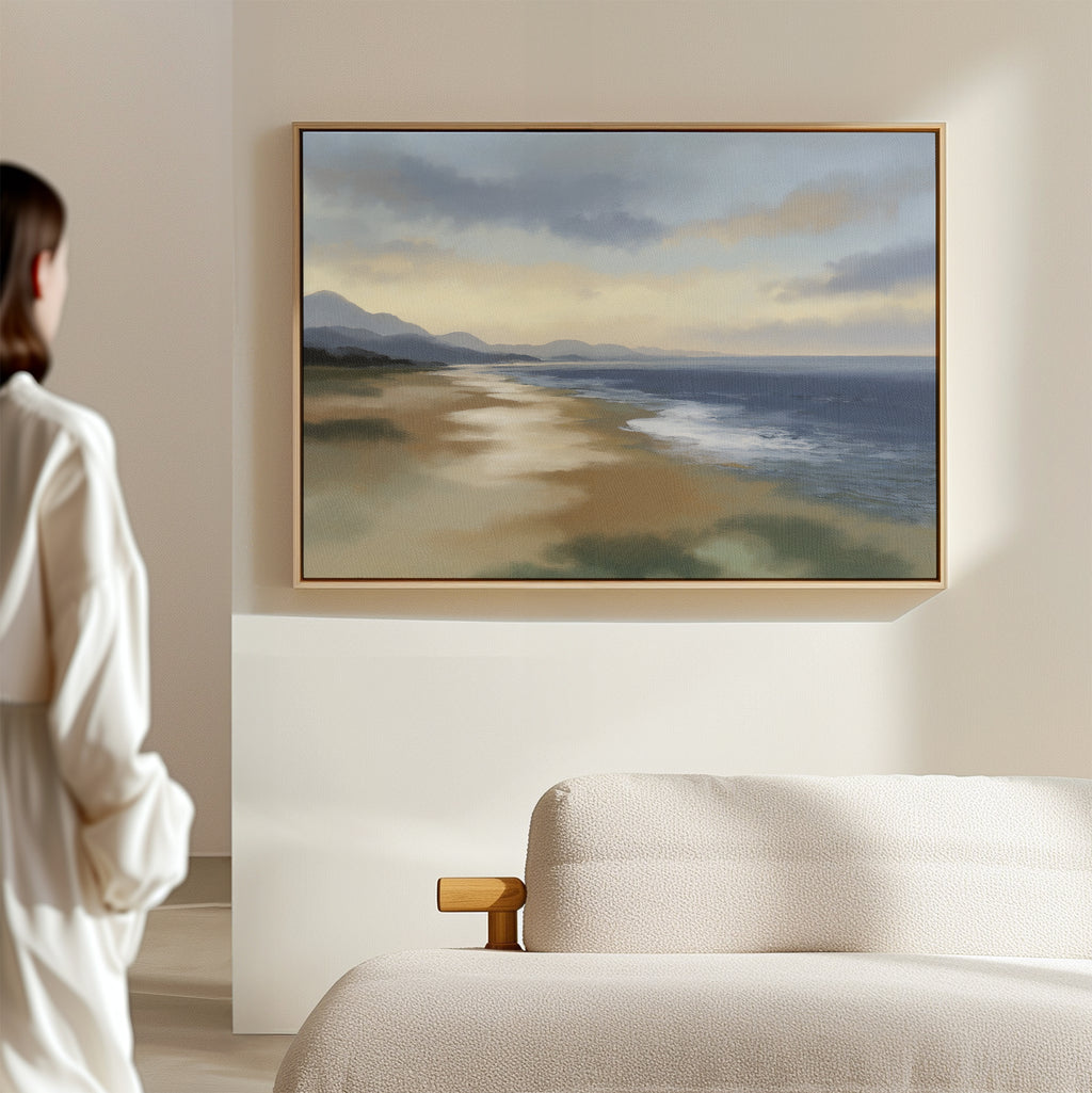 In a bright, minimal room, a person gazes at the Distant Shores Canvas Art - Horizontal, featuring gentle waves and distant mountains, displayed above a pale upholstered sofa.