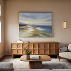 A cozy living room features a light wood coffee table, a round vase with flowers, a modern geometric sideboard, soft beige furnishings, and the "Distant Shores Canvas Art - Horizontal," bringing tranquil coastal vibes to the serene space.
