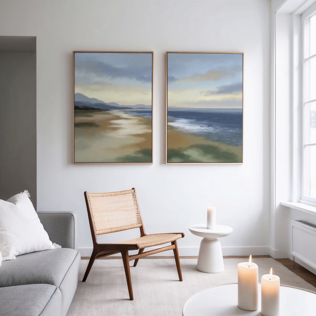 A bright, modern living room with neutral tones features a wooden chair, a round side table with a candle, a gray sofa, and Distant Shores - Set of 2 landscape canvas prints of a beach scene hung side by side on the white wall.