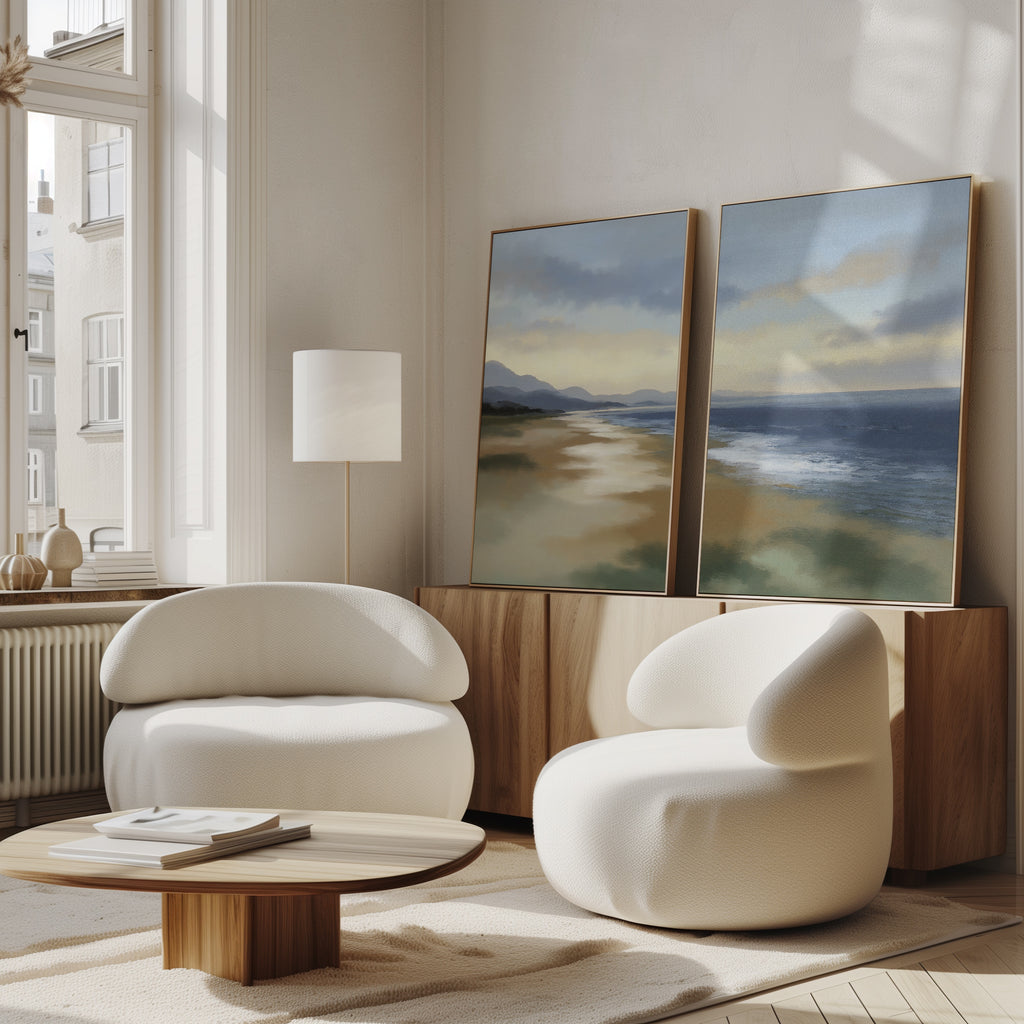A modern living room features two white, rounded armchairs, a low wooden coffee table, and a sideboard displaying the "Distant Shores - Set of 2" canvas prints. Serene coastal landscapes are illuminated by natural light streaming through tall windows.