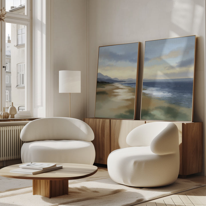 A modern living room features two white, rounded armchairs, a low wooden coffee table, and a sideboard displaying the "Distant Shores - Set of 2" canvas prints. Serene coastal landscapes are illuminated by natural light streaming through tall windows.