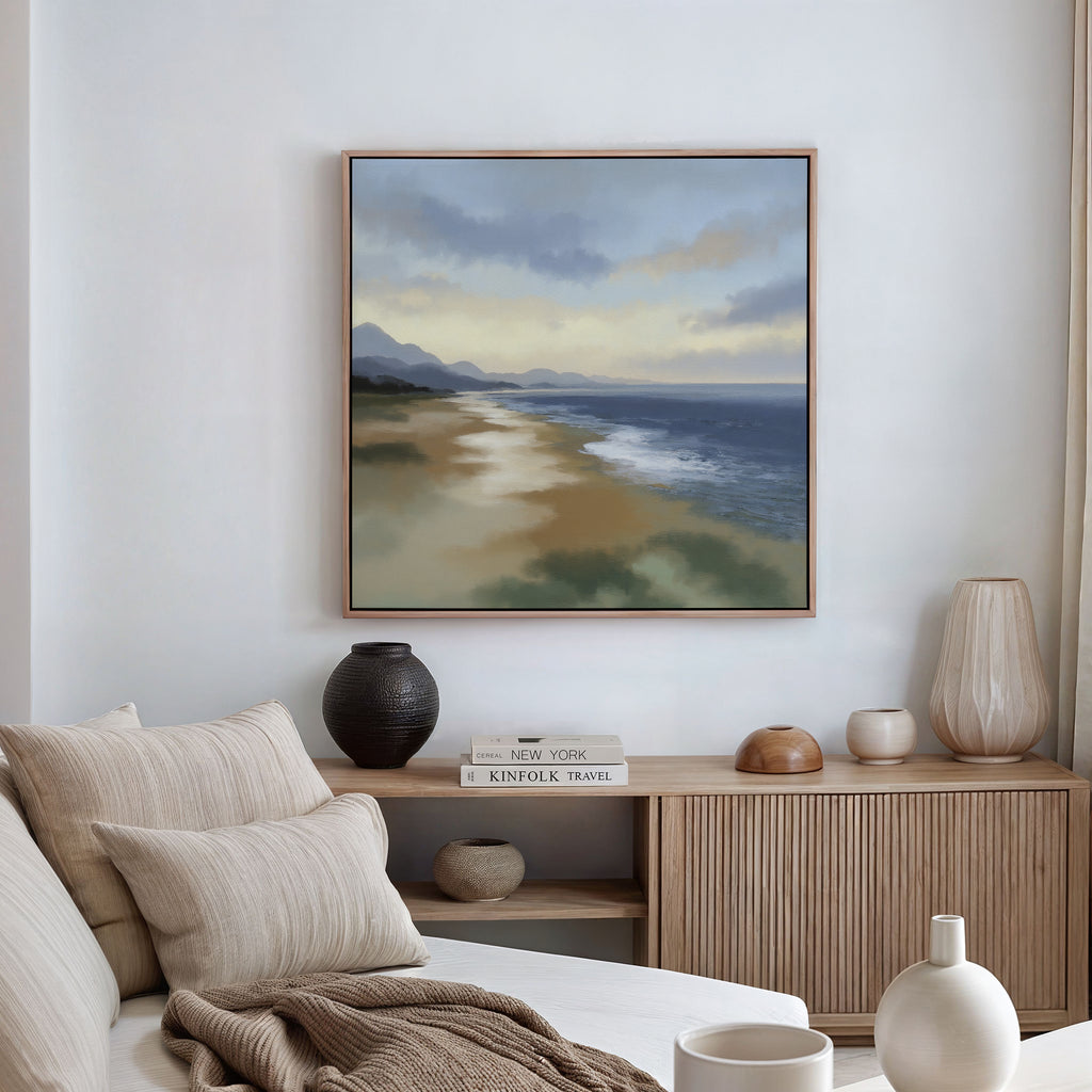 A cozy living room with a beige sofa, neutral cushions, a wooden sideboard with decor and books, and the Distant Shores Canvas Art - Square depicting a serene landscape hanging on the wall.