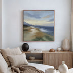 A cozy living room with a beige sofa, neutral cushions, a wooden sideboard with decor and books, and the Distant Shores Canvas Art - Square depicting a serene landscape hanging on the wall.