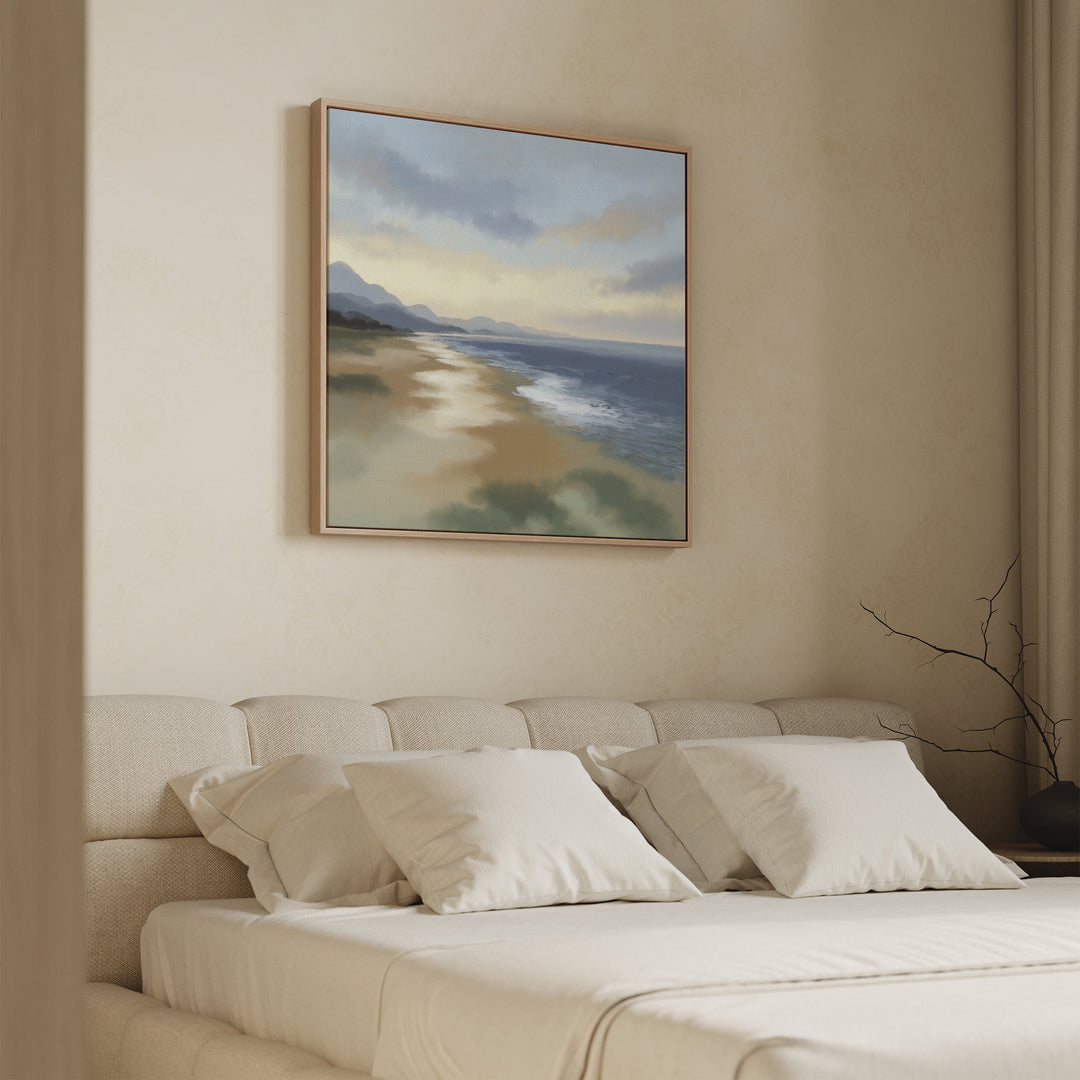 A serene bedroom with a beige upholstered bed, white pillows, and soft beige walls features the Distant Shores Canvas Art - Square above the bed, showcasing waves, sand, and distant hills beneath a cloudy sky.