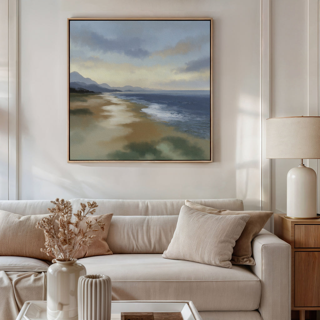 A cozy living room with a white sofa, beige cushions, a table with dried flowers, and a lamp. Above the sofa hangs the Distant Shores Canvas Art - Square, depicting waves, sand, and distant mountains under a cloudy sky.