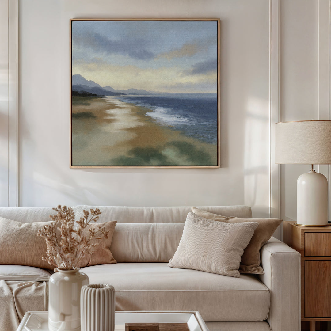 A cozy living room with a white sofa, beige cushions, a table with dried flowers, and a lamp. Above the sofa hangs the Distant Shores Canvas Art - Square, depicting waves, sand, and distant mountains under a cloudy sky.