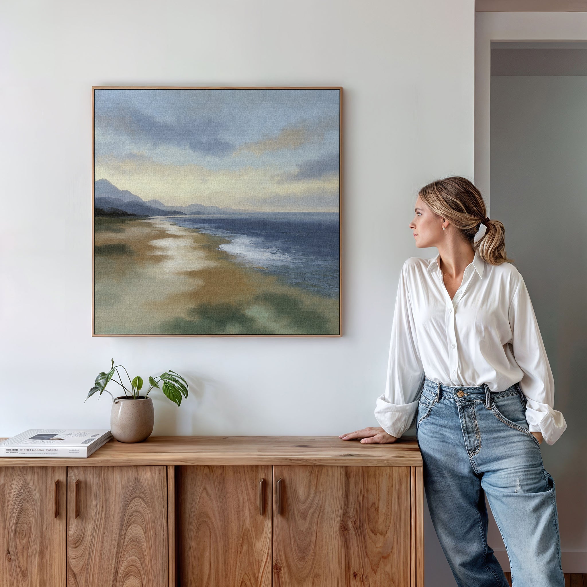 A woman in a white blouse and jeans stands by a wooden cabinet, admiring the Distant Shores Canvas Art - Square, which depicts a serene coastal panorama on the white wall behind her. A small potted plant and books sit atop the cabinet.