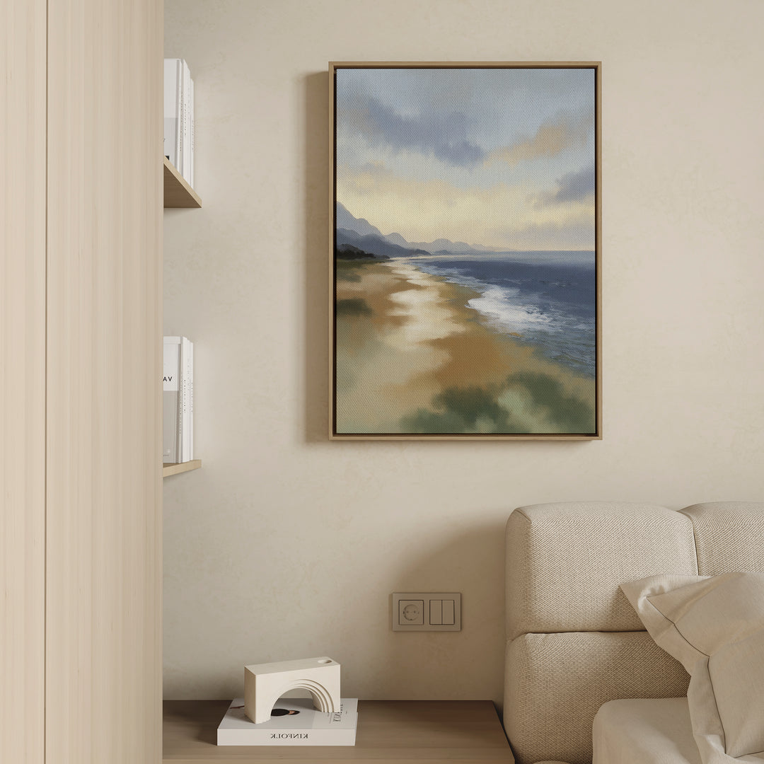 The Distant Shores Canvas Art - Vertical hangs on a beige wall above a light sofa, showcasing a serene shoreline with distant mountains and cloudy sky. Books and decor are arranged on shelves and a small table nearby.