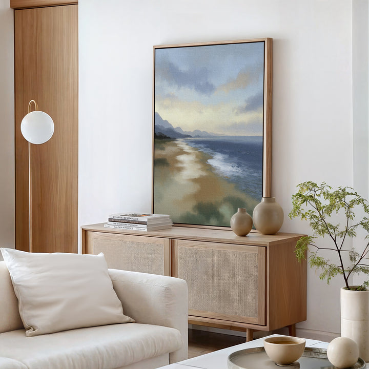 A modern living room featuring a light wood sideboard, the Distant Shores Canvas Art - Vertical, ceramic vases, books, a white sofa with a pillow, a small coffee table, a potted plant, and a round wall lamp.
