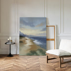 The Distant Shores Canvas Art - Vertical, featuring a coastal panorama with waves and mountains, leans against a white paneled wall in a stylish room with herringbone wood floors, a cushioned chair, round table, and candles.