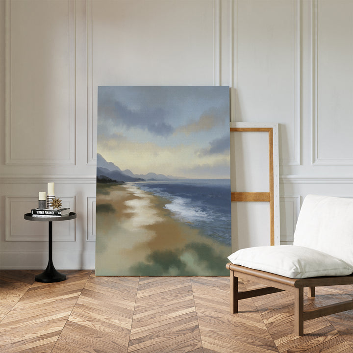 The Distant Shores Canvas Art - Vertical, featuring a coastal panorama with waves and mountains, leans against a white paneled wall in a stylish room with herringbone wood floors, a cushioned chair, round table, and candles.