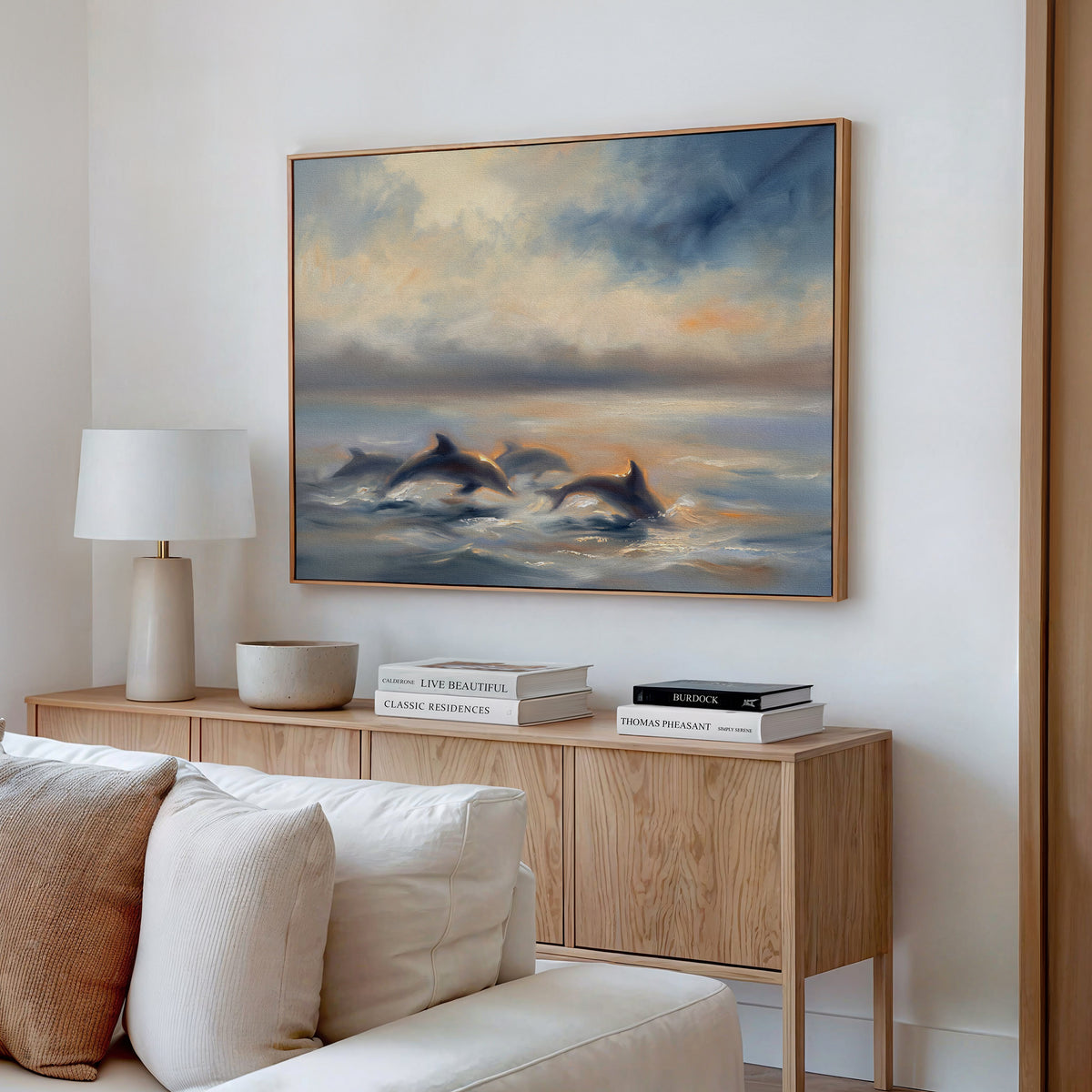 A cozy living room features the Dolphin Pod at Sunset Canvas Art - Horizontal swimming above a light wood sideboard with a white lamp, decorative bowl, and books. Neutral walls and a cream sofa enhance the tranquil, coastal-inspired decor.