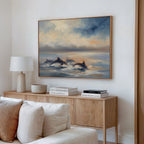 A cozy living room features the Dolphin Pod at Sunset Canvas Art - Horizontal swimming above a light wood sideboard with a white lamp, decorative bowl, and books. Neutral walls and a cream sofa enhance the tranquil, coastal-inspired decor.