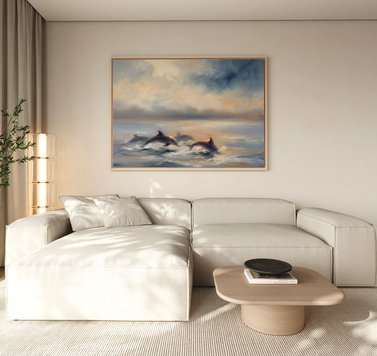 A modern living room features a cream sectional, light wood coffee table, and "Dolphin Pod at Sunset Canvas Art - Horizontal" on the beige wall. Natural light enters from the left.