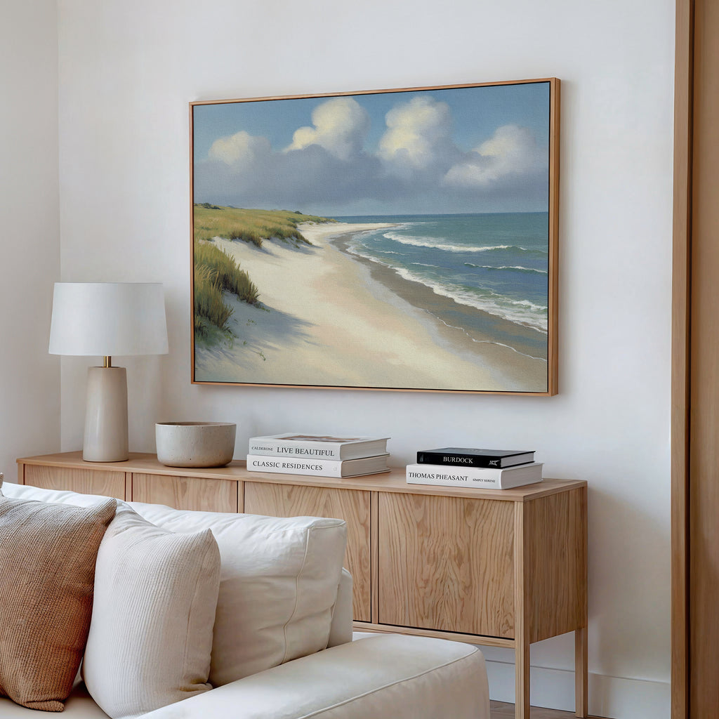 A cozy living room has a light wood sideboard with books, a lamp, and decor. Above it hangs the Dune Whispers Canvas Art - Horizontal—a serene coastal landscape. A soft beige sofa is placed in front.
