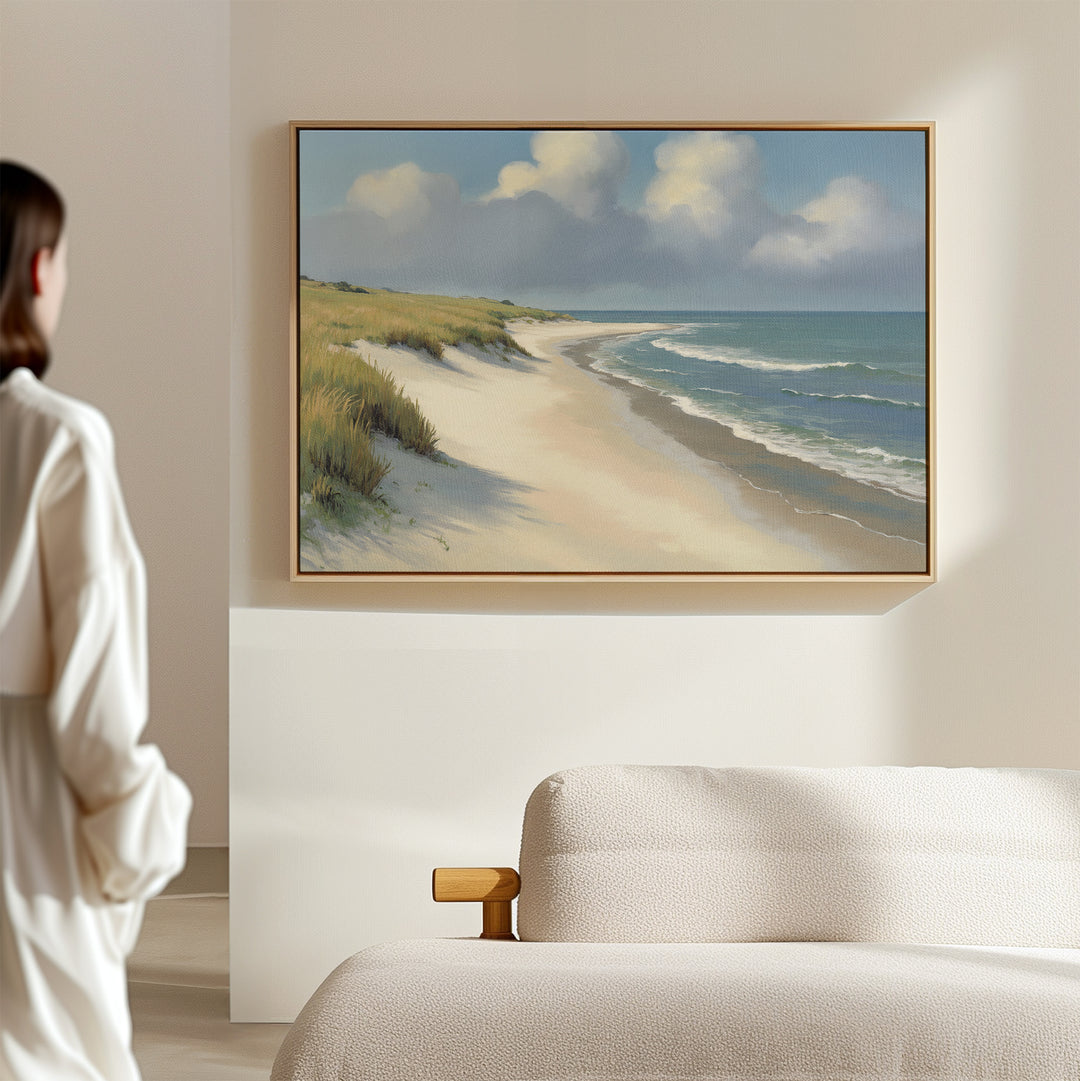 A person admires the Dune Whispers Canvas Art - Horizontal above a light sofa in a softly lit room. The artwork depicts a coastal landscape with sand, sea, grassy dunes, and a partly cloudy sky.