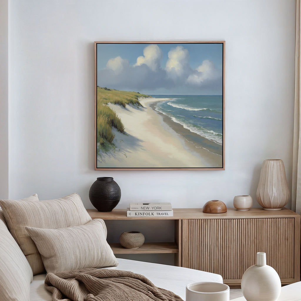 A coastal-themed living room features a beige sofa, neutral cushions, and a wooden sideboard. The wall displays the Dune Whispers Canvas Art - Square, showcasing sandy beach, ocean waves, grassy dunes, and clouds on a blue sky.