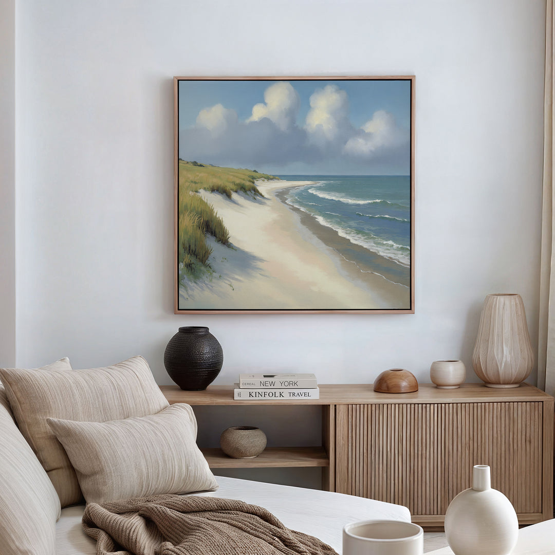 A coastal-themed living room features a beige sofa, neutral cushions, and a wooden sideboard. The wall displays the Dune Whispers Canvas Art - Square, showcasing sandy beach, ocean waves, grassy dunes, and clouds on a blue sky.
