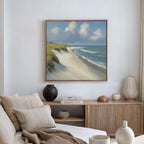 A coastal-themed living room features a beige sofa, neutral cushions, and a wooden sideboard. The wall displays the Dune Whispers Canvas Art - Square, showcasing sandy beach, ocean waves, grassy dunes, and clouds on a blue sky.