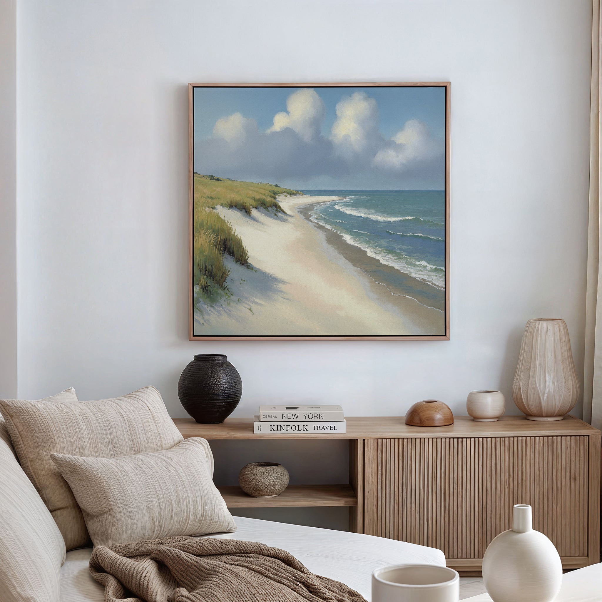 A coastal-themed living room features a beige sofa, neutral cushions, and a wooden sideboard. The wall displays the Dune Whispers Canvas Art - Square, showcasing sandy beach, ocean waves, grassy dunes, and clouds on a blue sky.
