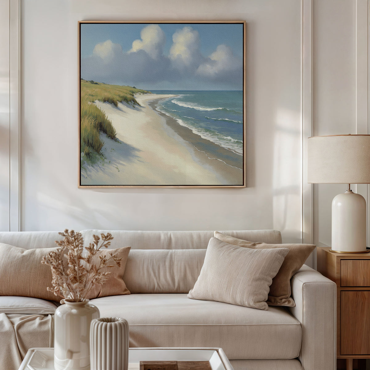 A cozy living room with a beige sofa, pillows, a side table and lamp, plus a vase of dried flowers. Above the sofa hangs the Dune Whispers Canvas Art - Square, depicting sand, grass, blue water, and a cloudy sky.
