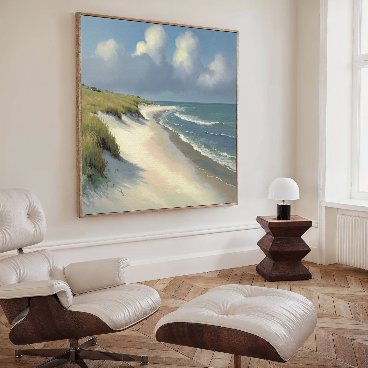 A modern minimalist living room features a cream lounge chair with ottoman, a wooden side table with white lamp, and the Dune Whispers Canvas Art - Square depicting sand dunes and ocean waves hanging on the wall.