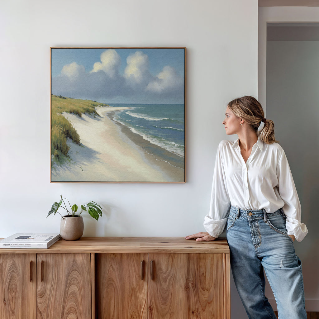 A woman in a white shirt and jeans stands by a wooden cabinet, gazing at the Dune Whispers Canvas Art - Square hanging on a white wall. A plant and a book rest on the cabinet.