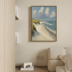 The Dune Whispers Canvas Art - Vertical, featuring a sandy beach with grass, waves, and clouds, hangs on a beige wall above a white sofa in a cozy modern living room filled with shelves and books.