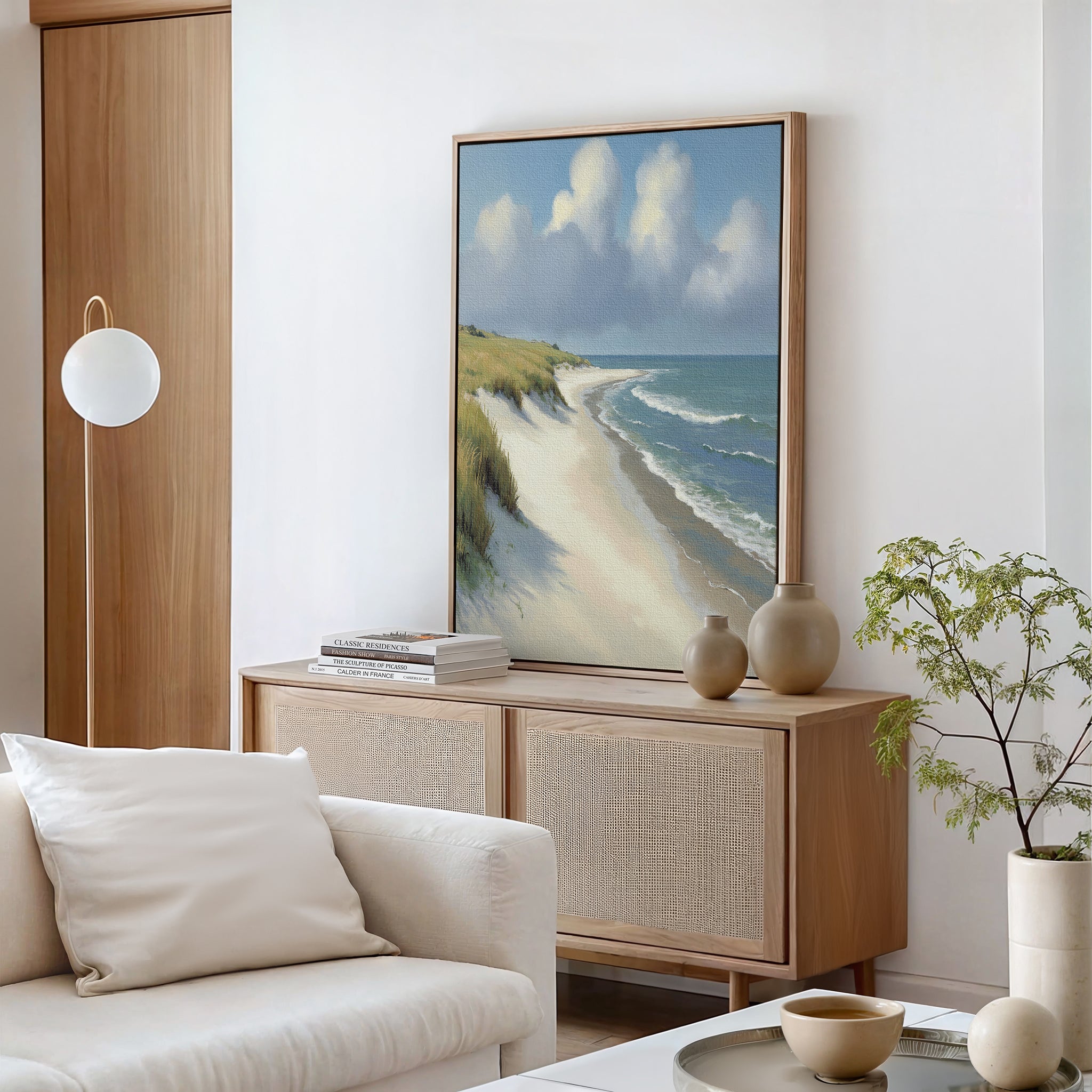 A modern living room features a light wood sideboard, Dune Whispers Canvas Art - Vertical, neutral ceramics, a white round sofa pillow, a small potted plant, and wooden accents against white walls.