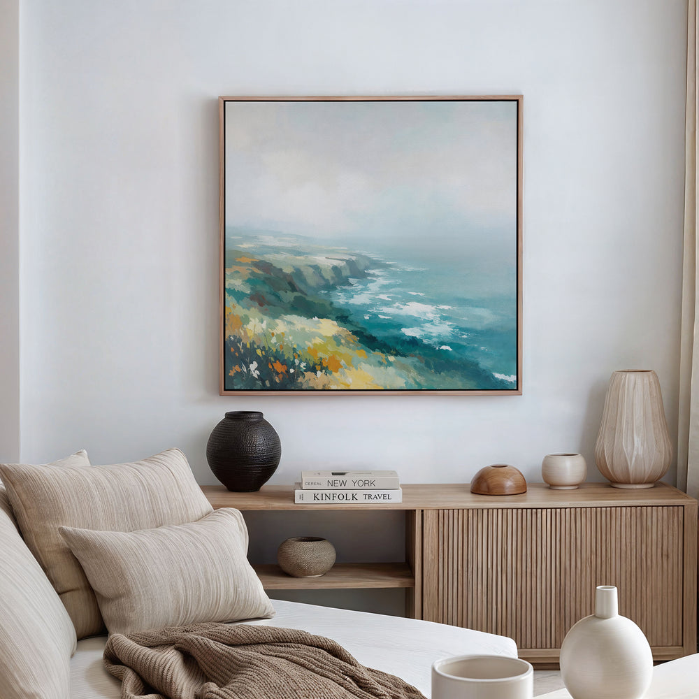 A modern living room featuring a beige sofa, neutral cushions, a wooden sideboard, ceramic vases, and the Emerald Cliffs Canvas Art - Square displaying a blue, green, and yellow seascape on a white wall.