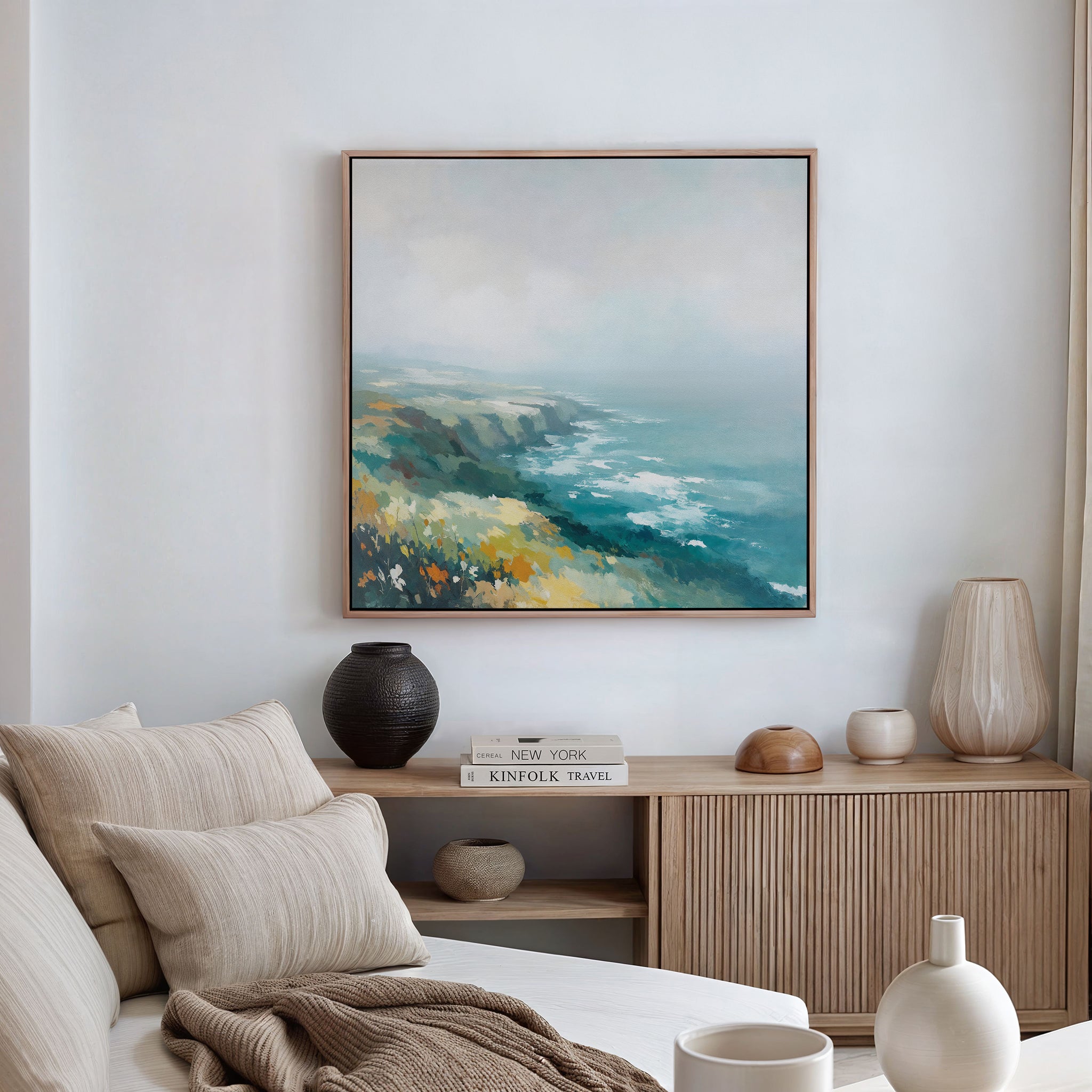 A modern living room featuring a beige sofa, neutral cushions, a wooden sideboard, ceramic vases, and the Emerald Cliffs Canvas Art - Square displaying a blue, green, and yellow seascape on a white wall.