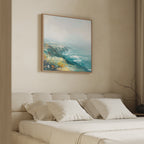 A minimal bedroom features a beige upholstered bed with white bedding and pillows. Above the bed hangs the Emerald Cliffs Canvas Art - Square, showcasing an abstract coastal landscape in blue, teal, yellow, and white.