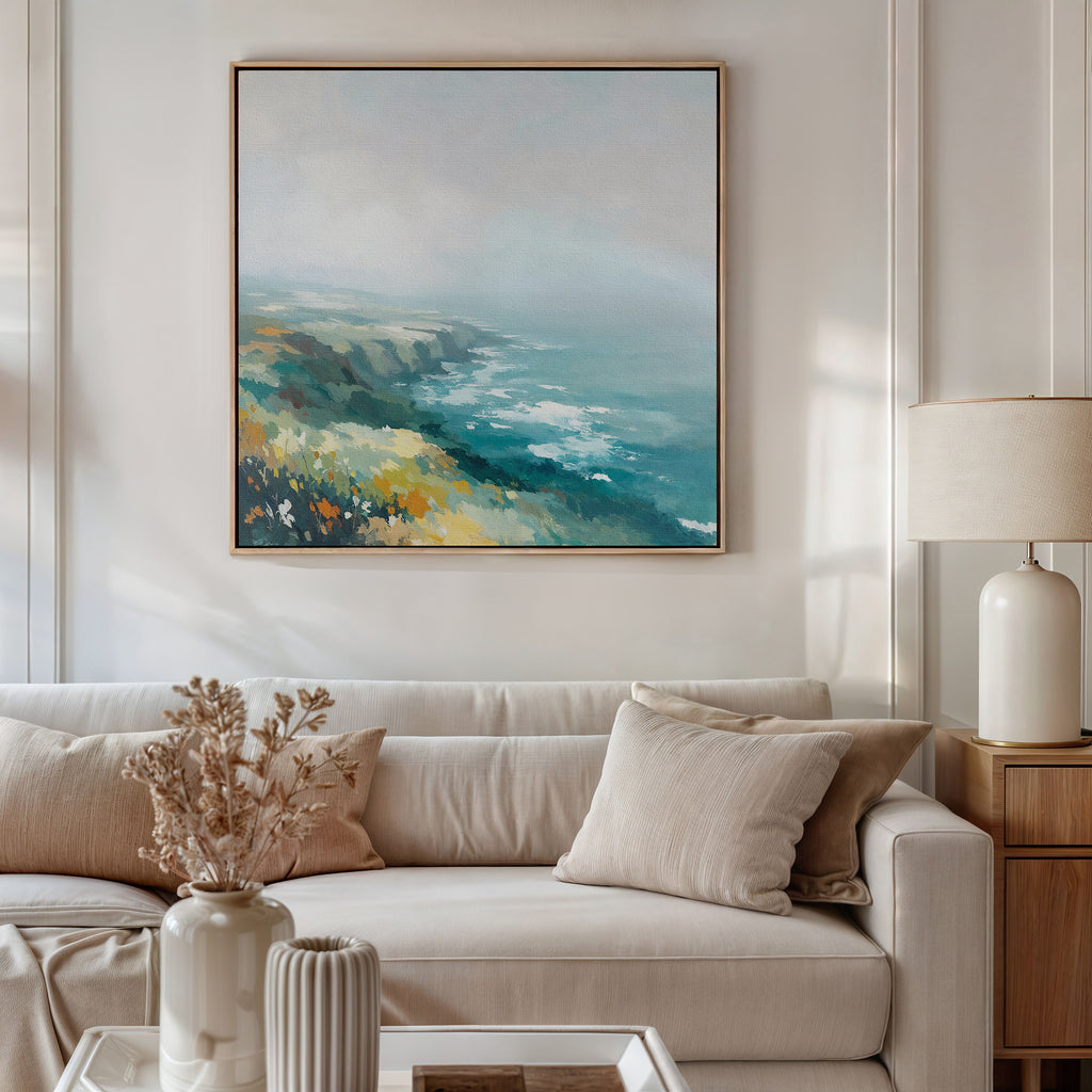 A modern living room features a beige sofa with cream cushions, a side table holding a lamp and dried flowers, and the Emerald Cliffs Canvas Art - Square hanging above, showcasing serene coastal beauty.