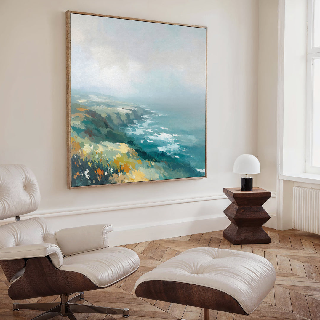 A modern, minimalist living room features a white leather lounge chair and ottoman, dark wood side table with white lamp, and a large square "Emerald Cliffs Canvas Art" coastal landscape on the wall, brightened by natural light from a tall window.