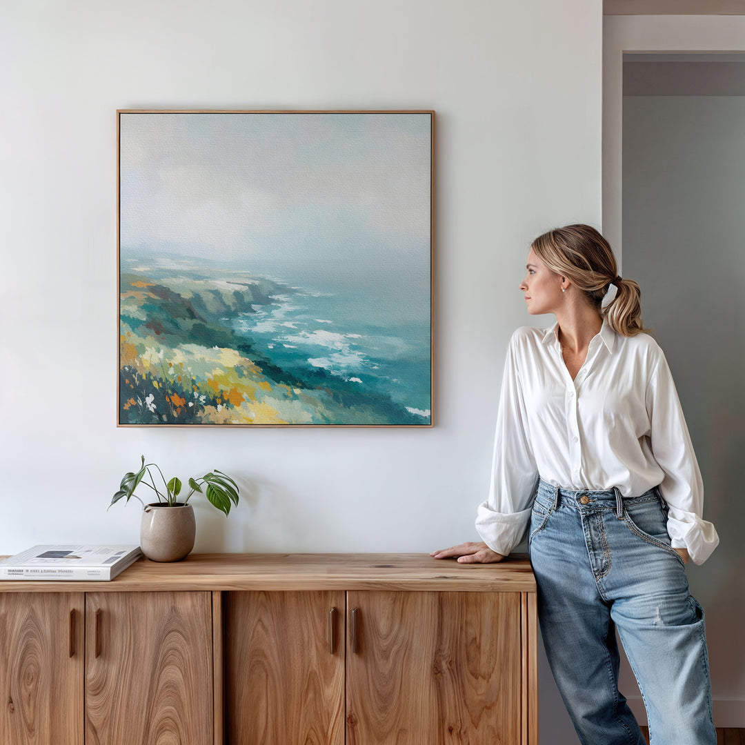 A woman in a white blouse and jeans stands by a wooden cabinet, gazing at the Emerald Cliffs Canvas Art - Square displayed above on a white wall. A small potted plant and books are neatly arranged on the cabinet beneath it.