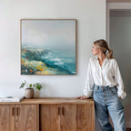 A woman in a white blouse and jeans stands by a wooden cabinet, gazing at the Emerald Cliffs Canvas Art - Square displayed above on a white wall. A small potted plant and books are neatly arranged on the cabinet beneath it.