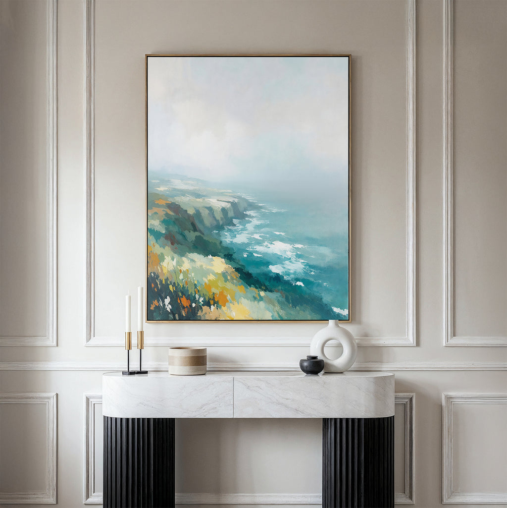 The Emerald Cliffs Canvas Art - Vertical hangs on a beige wall above a marble console table decorated with modern vases and candles. Elegant, classic wall molding enhances the coastal landscape’s foggy ocean and cliff scene.