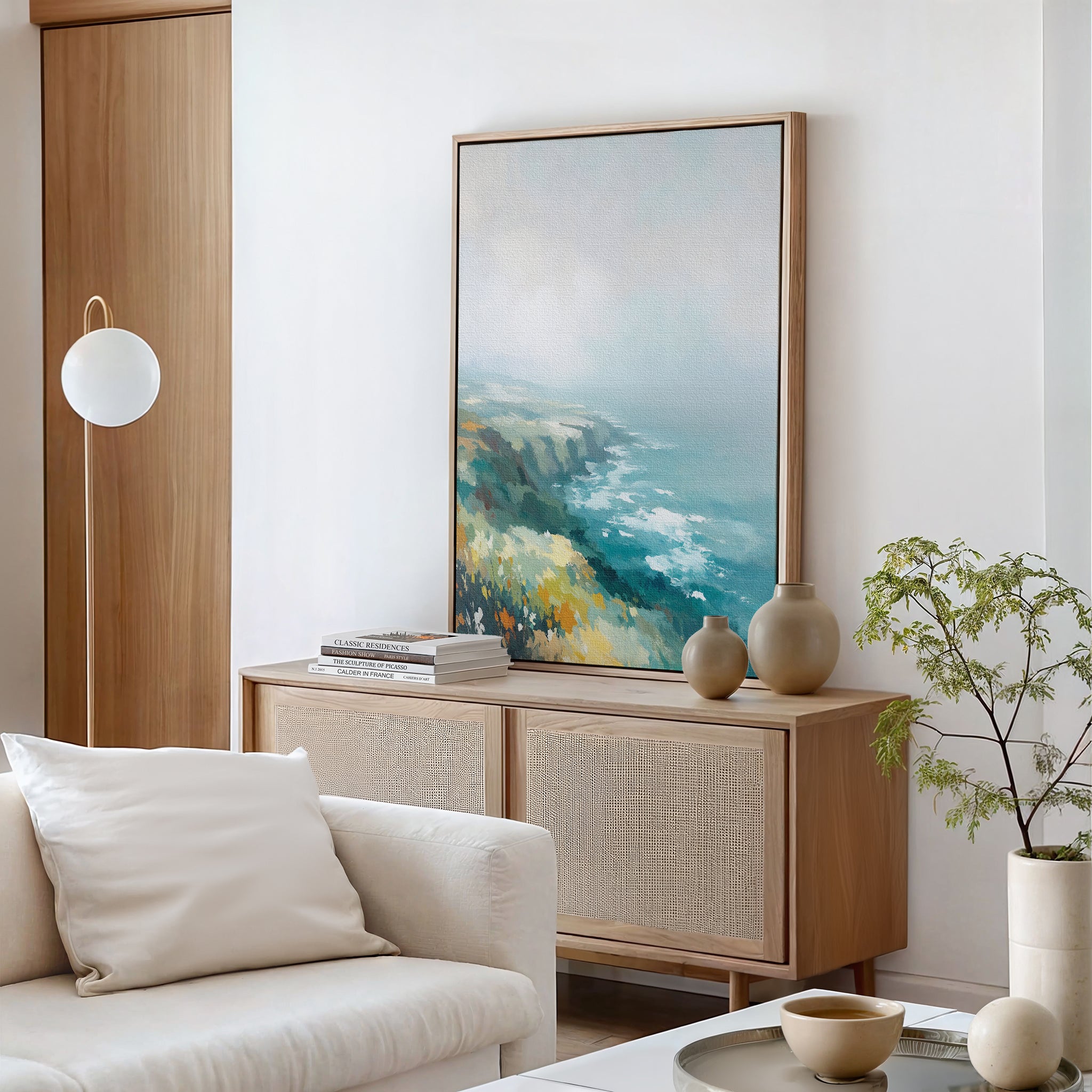 A modern living room featuring a light wood sideboard, neutral decor, the vertical Emerald Cliffs Canvas Art, potted plant, beige sofa with cushions, books, ceramic vases, and a round white pendant lamp.