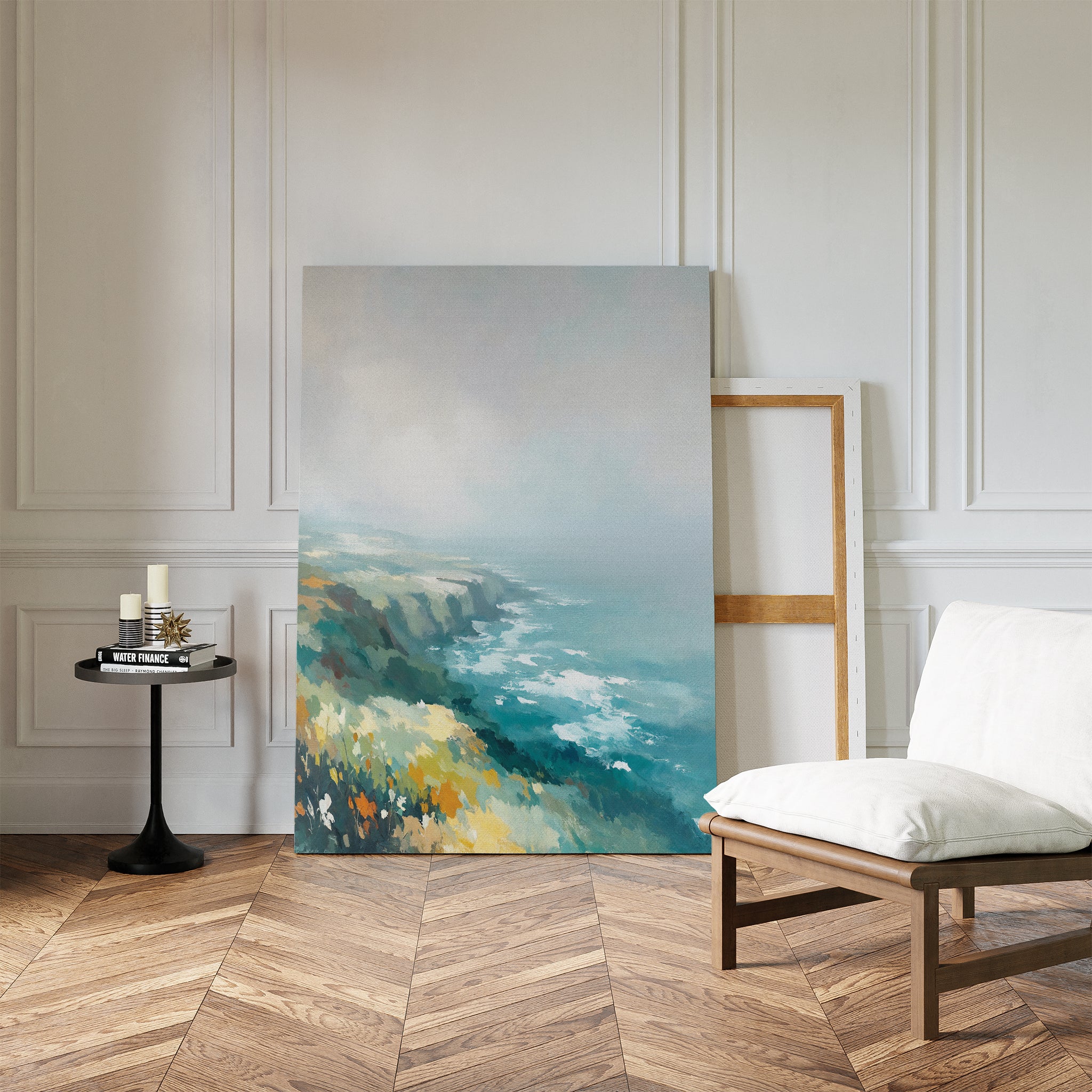 The Emerald Cliffs Canvas Art - Vertical, featuring a pastel-hued misty coastline, leans against a white wall next to an empty wooden frame in a modern room with herringbone floors, a cushioned chair, and a black round table with candles.