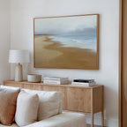 A modern living room features a light wood sideboard with books, a lamp, and bowl. Above the cream sofa hangs serene wall art—a framed Endless Shoreline Canvas Art - Horizontal—on the white wall.