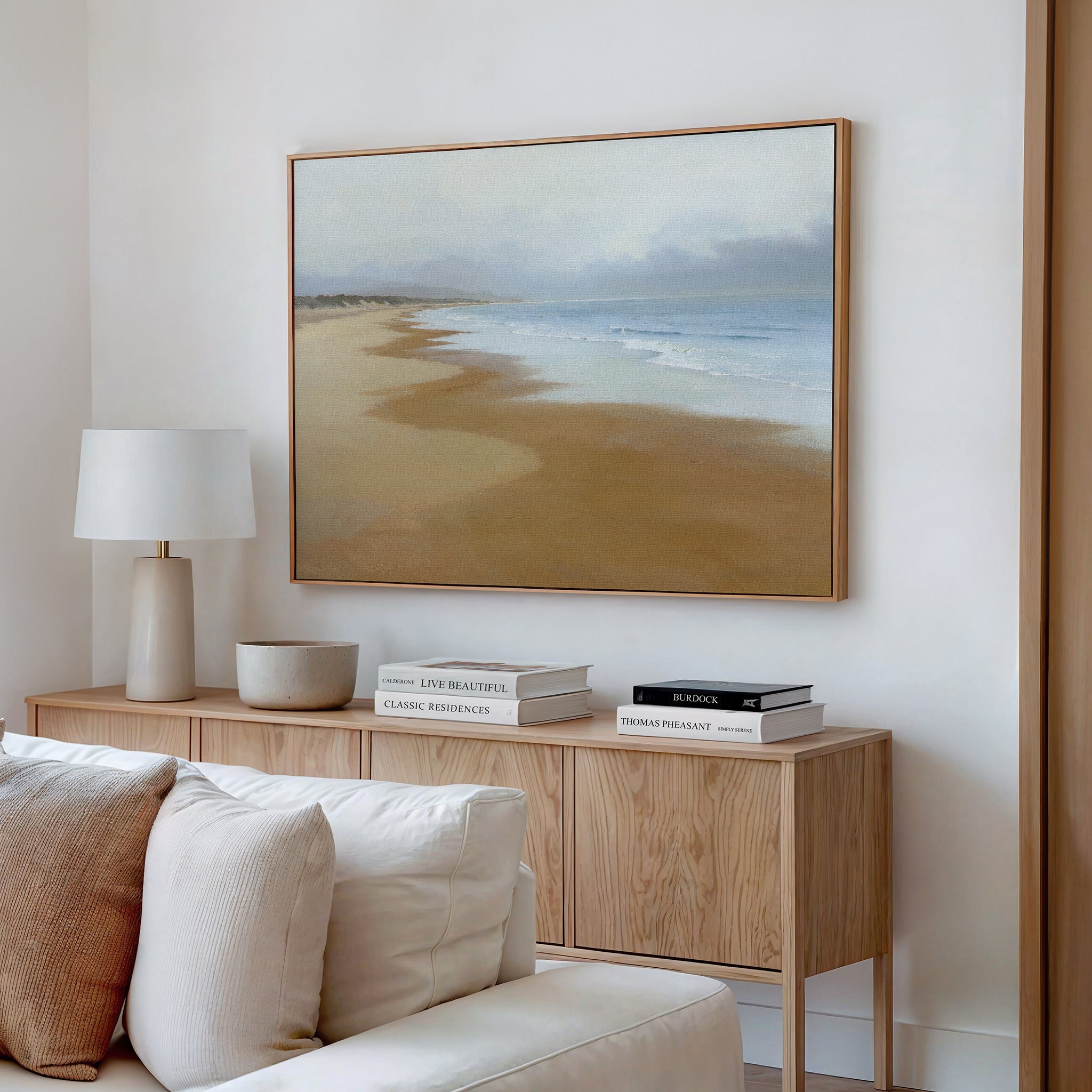 A modern living room features a light wood sideboard with books, a lamp, and bowl. Above the cream sofa hangs serene wall art—a framed Endless Shoreline Canvas Art - Horizontal—on the white wall.