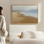 A woman in a white robe stands in a minimalist room, gazing at the Endless Shoreline Canvas Art - Horizontal, featuring sand, sea, and sky, displayed above a beige sofa on a light-colored wall.
