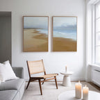 A modern living room with a gray sofa, wicker chair, small white side table, and three lit candles. "Endless Shoreline - Set of 2" serene coastal wall art hangs above the chair. Natural light brightens the Nordic-inspired space.