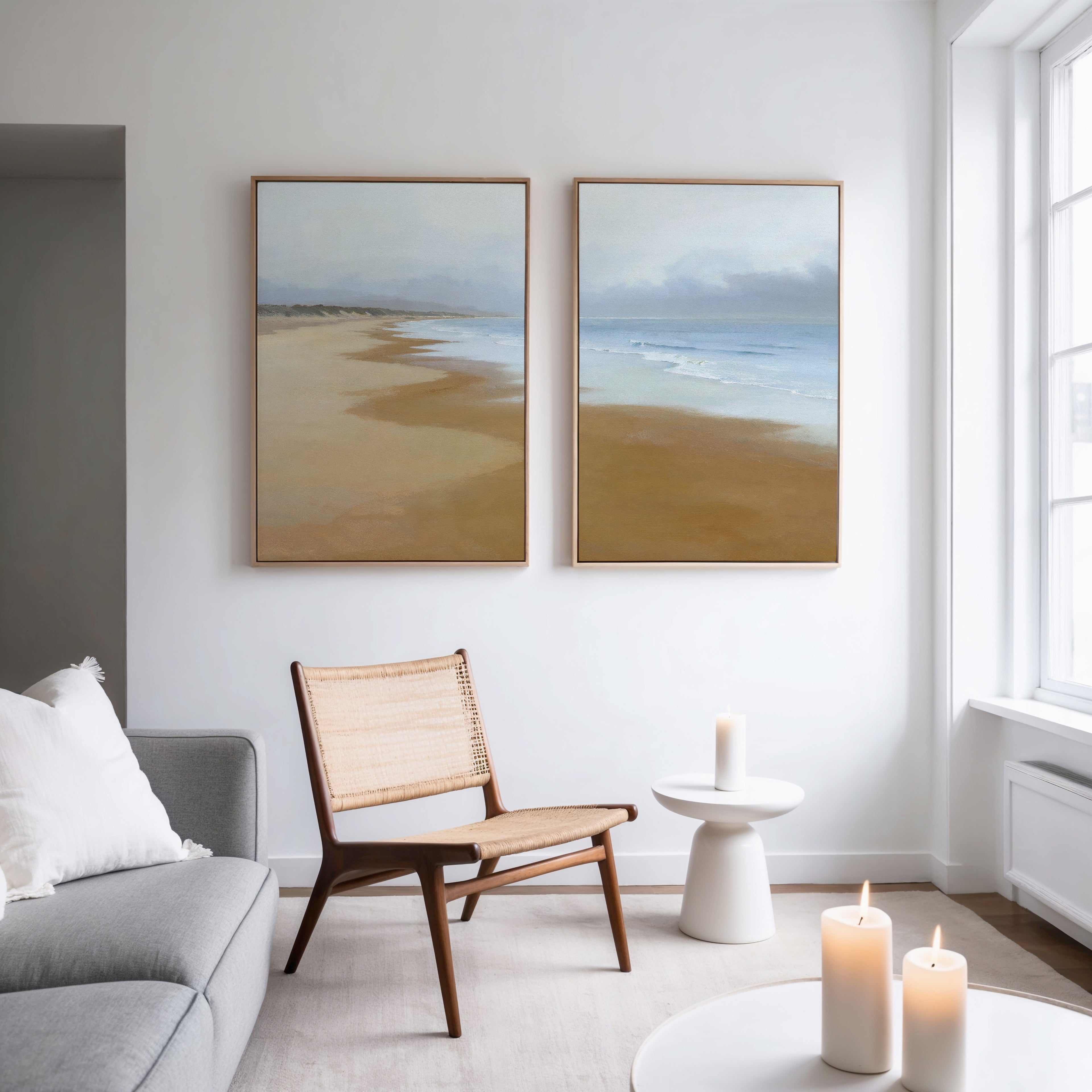 A modern living room with a gray sofa, wicker chair, small white side table, and three lit candles. "Endless Shoreline - Set of 2" serene coastal wall art hangs above the chair. Natural light brightens the Nordic-inspired space.