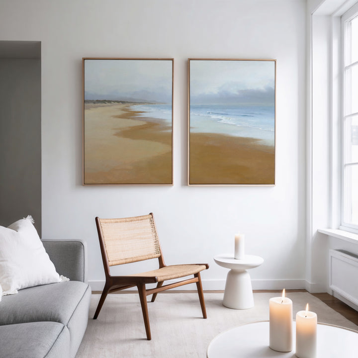 A modern living room with a gray sofa, wicker chair, small white side table, and three lit candles. "Endless Shoreline - Set of 2" serene coastal wall art hangs above the chair. Natural light brightens the Nordic-inspired space.